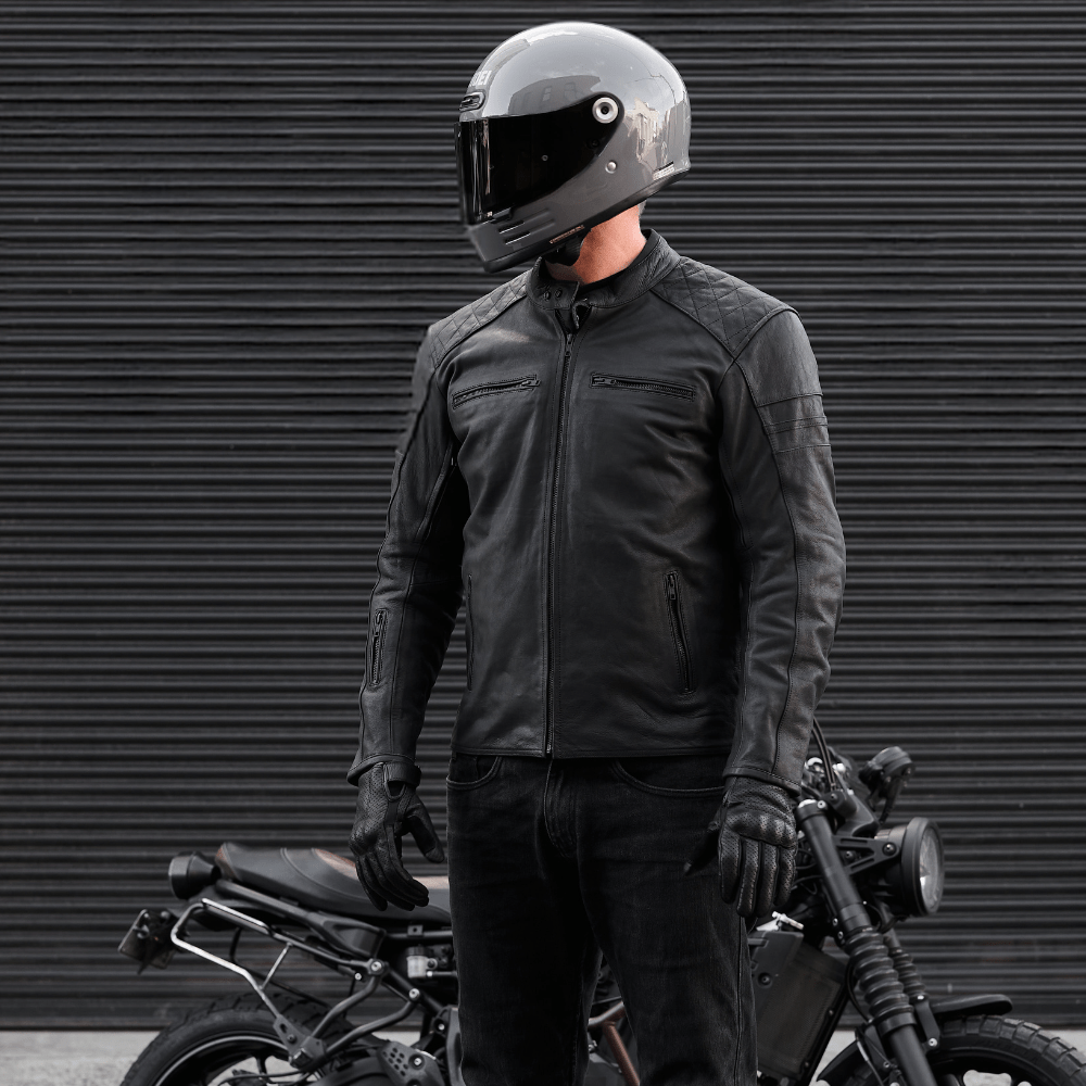 Person wearing a black motorcycle helmet and jacket standing next to a motorcycle against a dark background