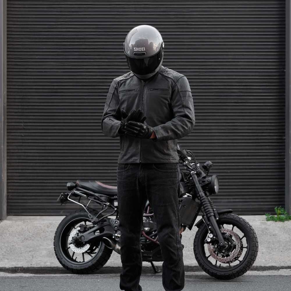 Person wearing a black helmet and jacket standing next to a motorcycle in front of a gray wall.