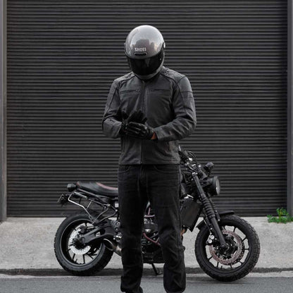 Person wearing a black helmet and jacket standing next to a motorcycle in front of a gray wall.