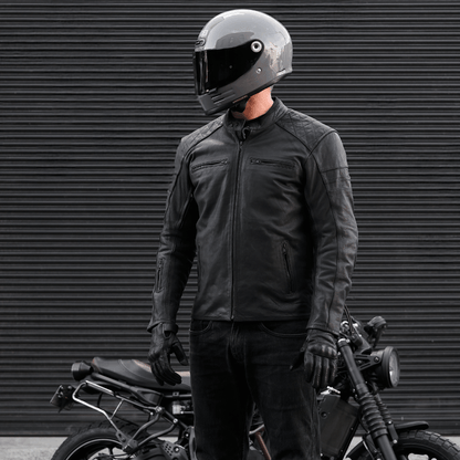 Person wearing a black motorcycle helmet and jacket standing next to a motorcycle against a dark background