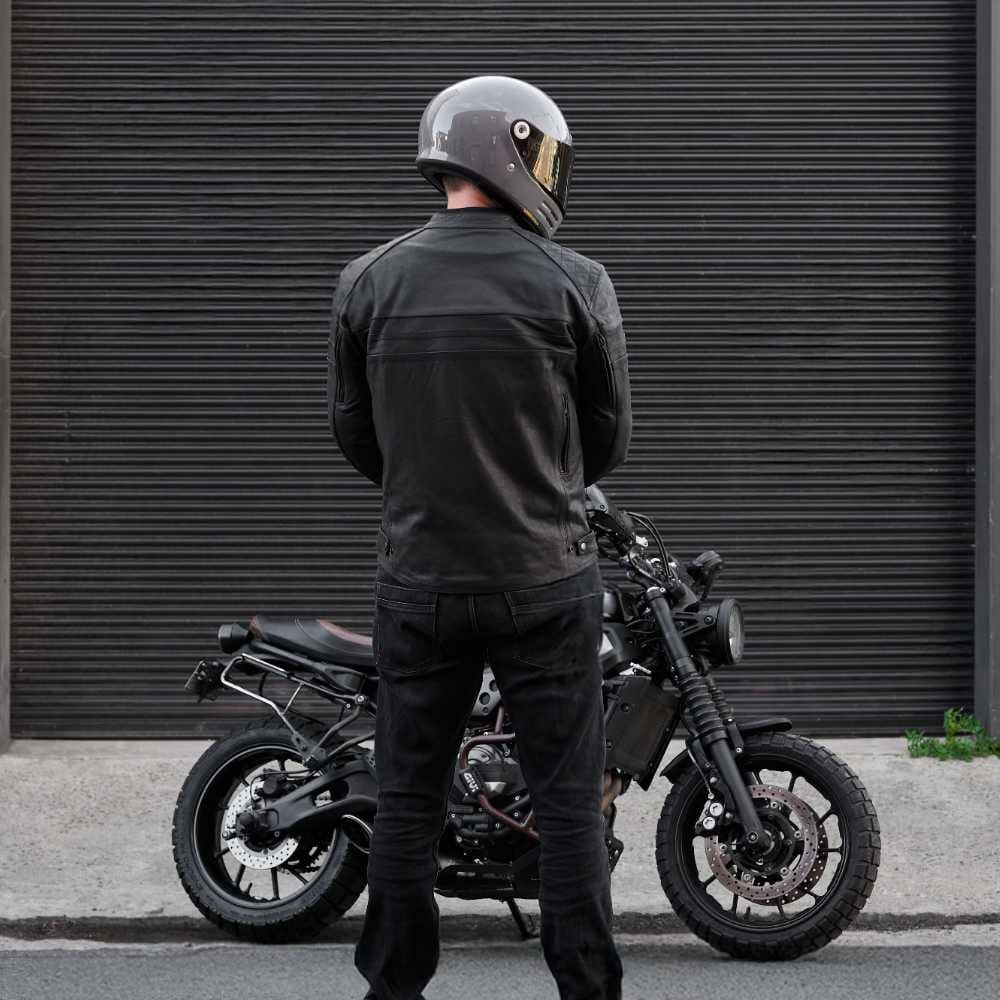 Person wearing a helmet and dark clothing standing next to a motorcycle against a dark background