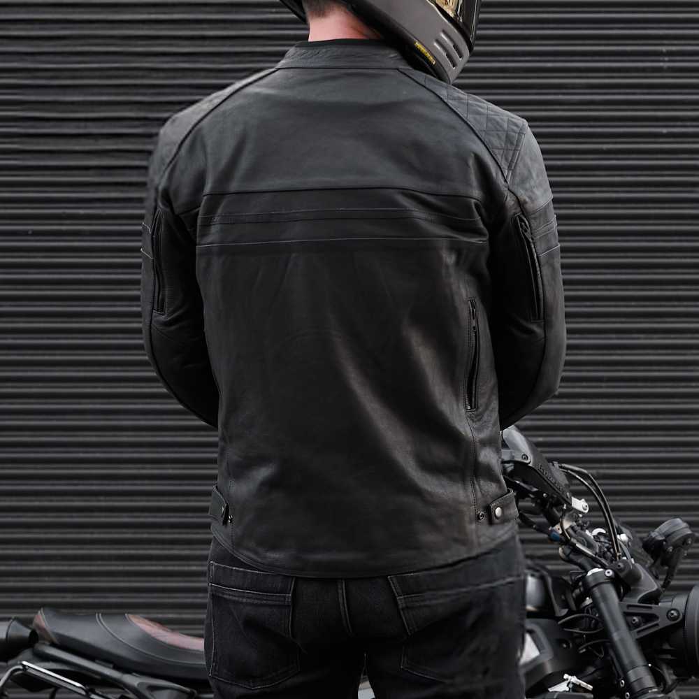 Person wearing a black motorcycle jacket standing next to a motorcycle with a dark background